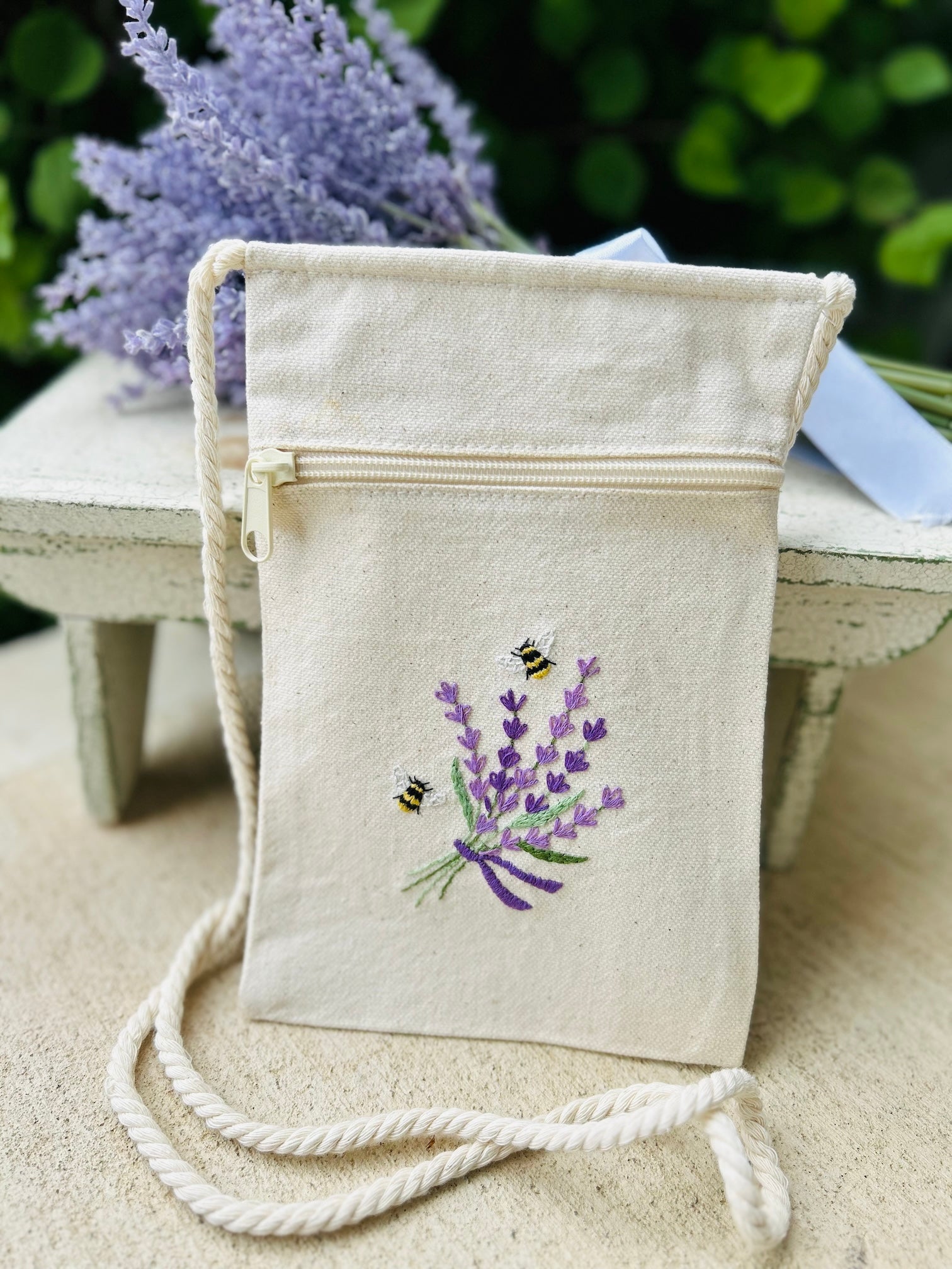 A beige cross-body canvas bag with a twisted rope strap, featuring an embroidered design of bees and lavender, designed by Deb Strain.