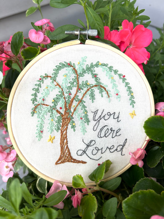 An embroidery kit depicting a willow tree with the phrase 'You Are Loved' displayed in a hoop, designed by Deb Strain.
